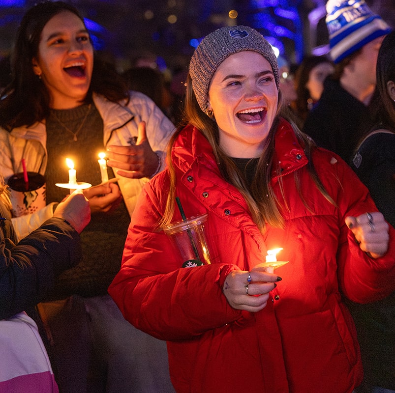 Two students laugh while wearing winter coats and holding candles.