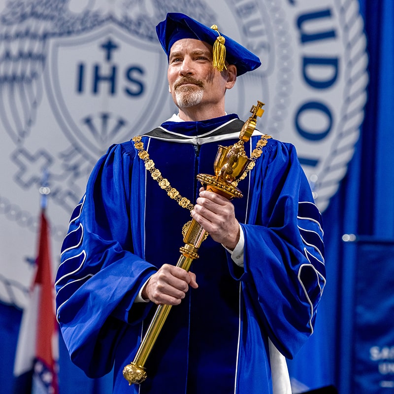 President Feser holds the presidential mace while wearing an academic robe and cap.