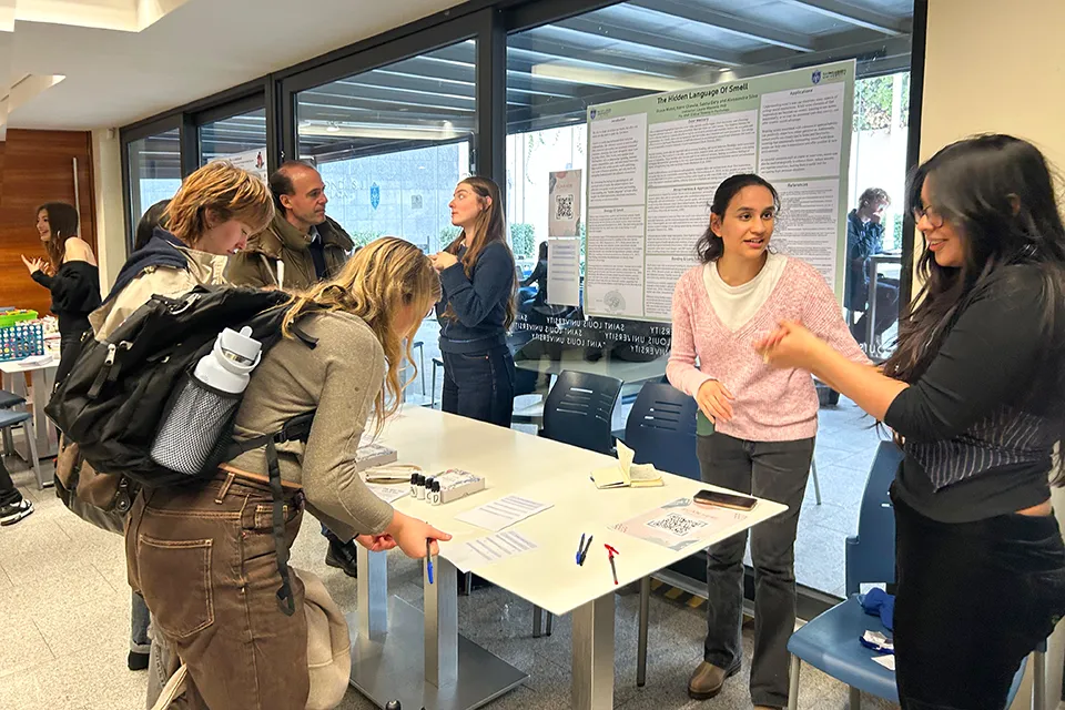 SLU-Madrid Psychology Majors Share Projects with Peers Two people look down while filling out a form while looking at essential oil bottles. Two girls talk to event participants behind a table and a poster in the background shows research details.