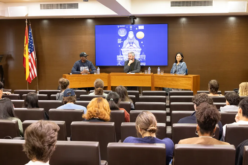 Best-selling Fantasy Authors Lead Creative Writing Event at SLU-Madrid A panel of three speakers sits at a wooden table in front of an audience in a lecture hall. Behind them is a large screen displaying event artwork. The speaker on the left wears a cap and sweatshirt, the middle speaker rests his hand on his chin, and the speaker on the right smiles.