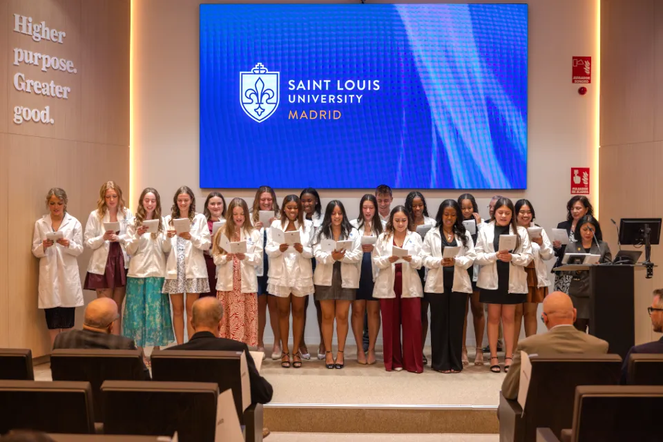 SLU-Madrid Marks 11th Annual White Coat Ceremony Students with white coats on are reading off pamphlets together in front of an audience. The logo of the University is on the screen behind them. A sign on the wall reads "Higher purpose. Greater good."