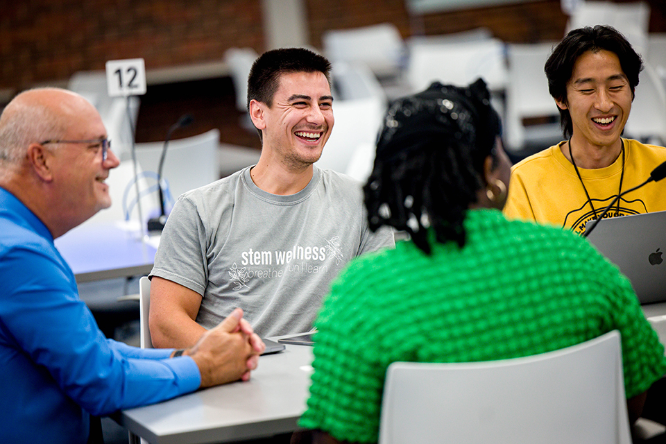 Anthony Breitbach, Ph.D., sits a table with two male students wearing gray and yellow shirts and one female student wearing a green shirt during an interprofessional class on Oct. 2, 2023. The group are smiling. 