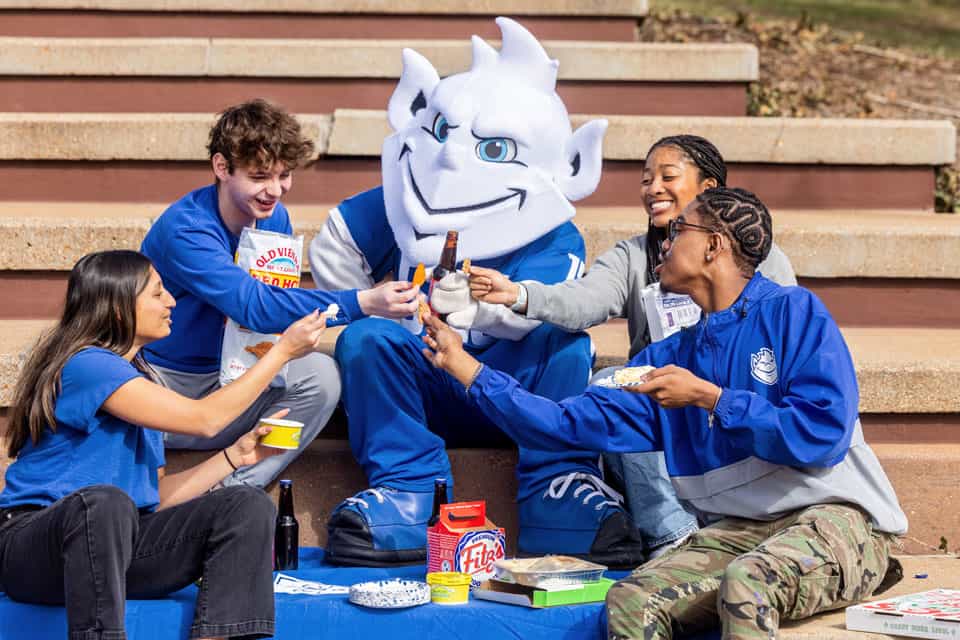 Four students surrounding the billiken mascot with local st louis foods on steps by SLU clocktower